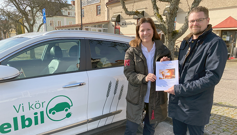 En kvinna och en man står framför en vit bil. Båda håller i ett diplom.