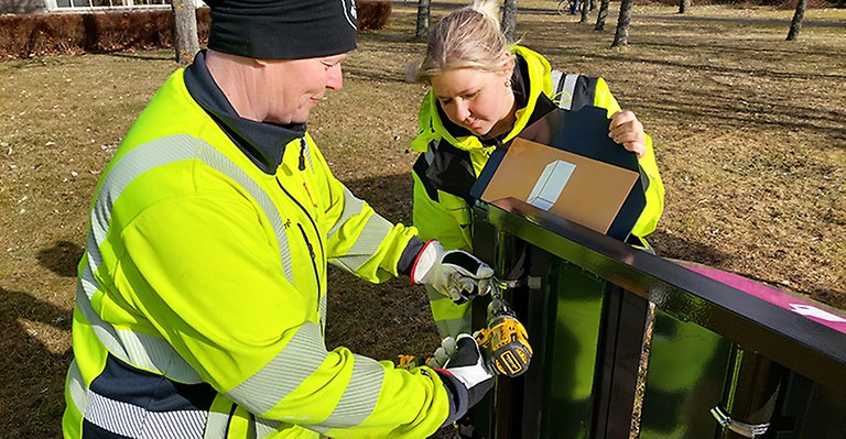 Två kvinnor i varselkläder monterar upp en behållare på en ställning.